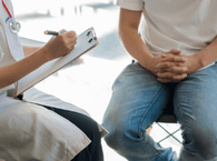 A patient sits with a medical professional who is taking notes on a clipboard.