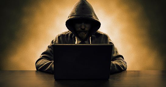 A hooded figure sits at a laptop, shrouded in darkness.