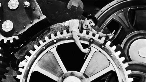 A movie still featuring Charlie Chaplin laying on a large piece of machinery.