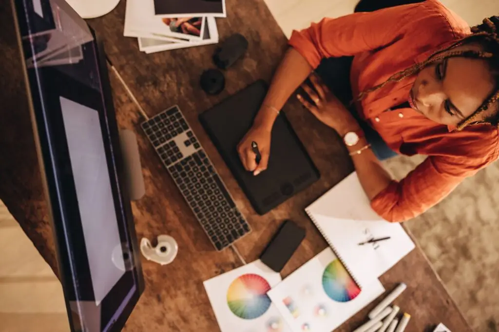 Overhead shot of a woman using a pen tablet, with a monitor, keyboard, color wheels, and markers on a wooden desk.