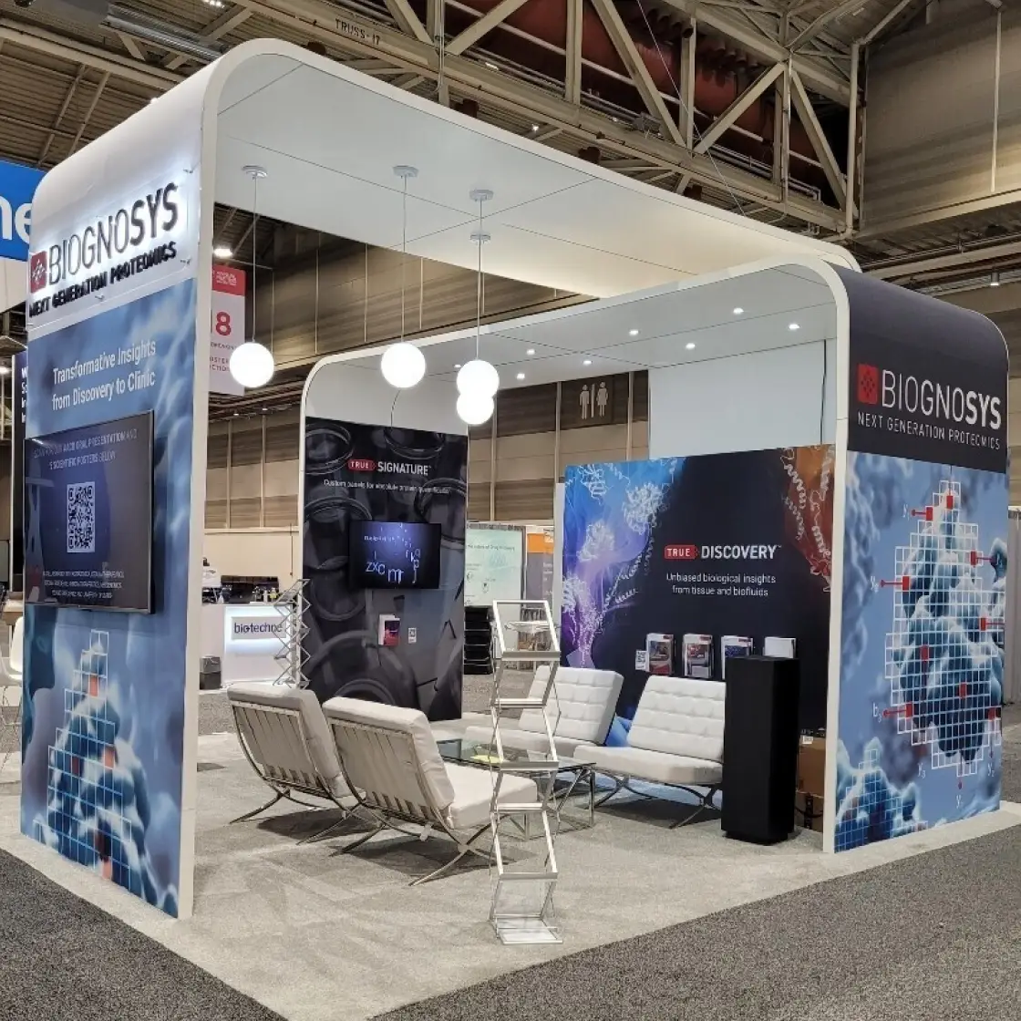 A Biognosys booth with "Next Generation Proteomics" and "Transformative Insights from Discovery to Clinic" text, featuring screens and seating.