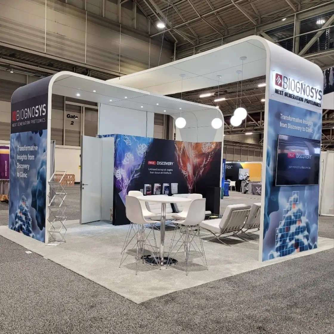 A Biognosys booth with "Next Generation Proteomics" and "Transformative Insights from Discovery to Clinic" displayed, featuring a screen and seating.