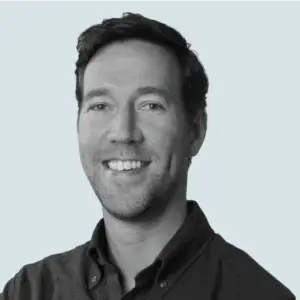 John Pantlind, VP, Media headshot: A black and white headshot of a smiling man with short dark hair and a dark collared shirt.