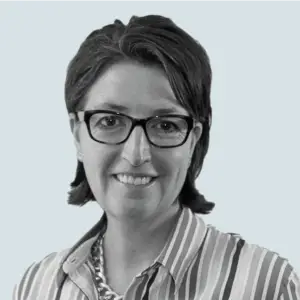 Rebecca Angelos, Chief Operating Officer headshot: A black and white headshot of a smiling woman with short dark hair and glasses, wearing a striped shirt and a chunky necklace.