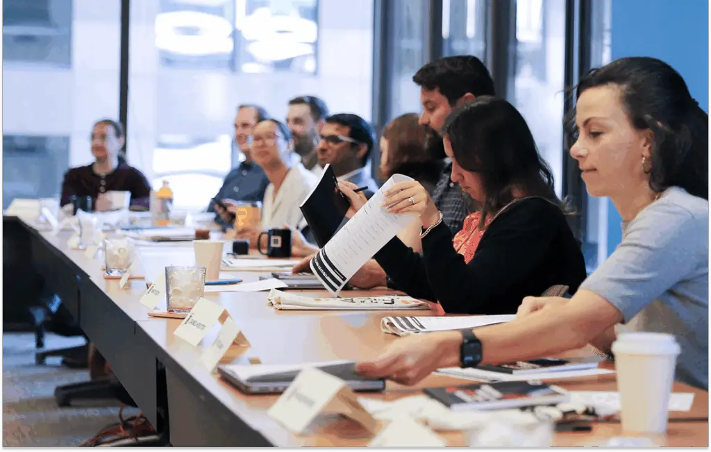 A diverse group of professionals sits around a conference table, engaged in a meeting. Some are looking at documents, while others listen attentively.