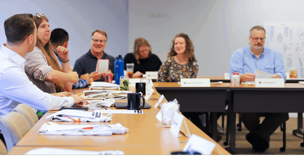 A group of people sitting around a conference table, engaged in a meeting or discussion. Some are looking at papers, others are listening.